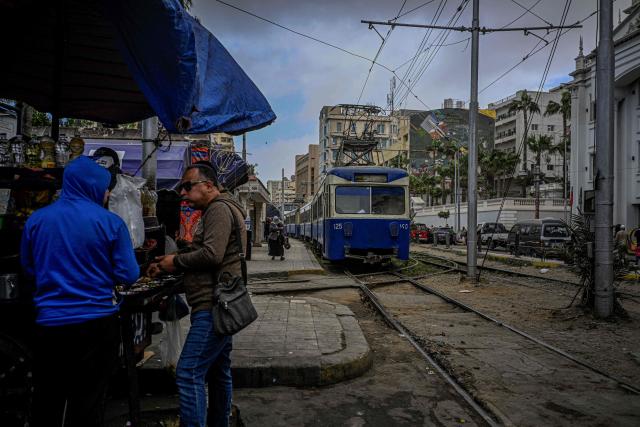 Egyptians stand at a food stall set up along the tram tracks in the coastal city of Alexandria on February 18, 2026. Along Egypt's Mediterranean coast, the oldest tram in Africa and the Middle East rumbles for a final few weeks before its removal -- the latest urban upheaval Alexandrians say is hollowing out their city's identity. Inaugurated in 1863, the tram is one of the world's oldest, and among only a few to operate double-decker cars. (Photo by Khaled DESOUKI / AFP)
