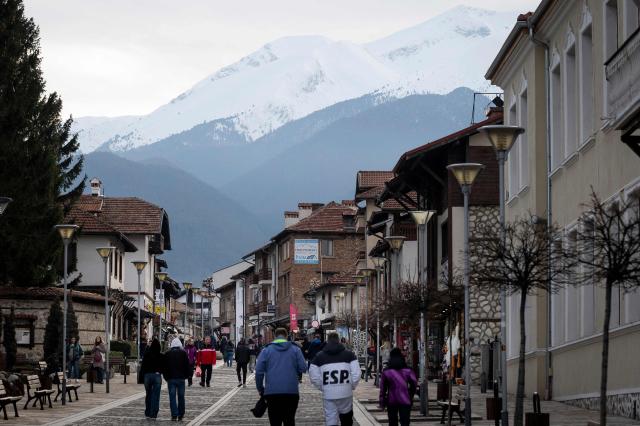Pedestrians walk along a street in the centre of Bansko with Pirin Mountain in the background on February 14, 2026. Bulgaria's largest ski resort of Bansko in Pirin mountain is becoming a hub for digital nomads, copywriters, enterpreneurs and retirees from around the world. (Photo by Nikolay DOYCHINOV / AFP) / TO GO WITH AFP STORY BY ROSSEN BOSSEV