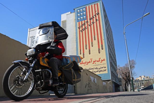 TOPSHOT - An Iranian man rides past an anti-US mural on a building in Tehran on February 26, 2026. A third round of indirect talks between the United States and Iran, mediated by Oman's foreign minister, opened in Geneva on February 26 morning, a diplomatic source told AFP. (Photo by AFP)