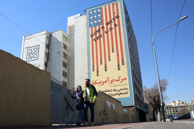An Iranian couple walk past an anti-US mural on a building in Tehran on February 26, 2026. A third round of indirect talks between the United States and Iran, mediated by Oman's foreign minister, opened in Geneva on February 26 morning, a diplomatic source told AFP. (Photo by AFP)