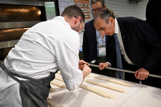 French far-right Reconquete party leader Eric Zemmour (R) observes how to make an incision in a baguette at a baker's booth during his visit of the Paris International Agricultural Show (Salon de l'Agriculture) at Paris Expo Porte de Versailles in Paris on February 26, 2026. (Photo by Anna KURTH / AFP)