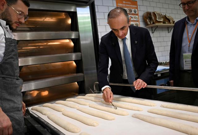 French far-right Reconquete party leader Eric Zemmour (C) makes an incision in a baguette at a baker's booth during his visit of the Paris International Agricultural Show (Salon de l'Agriculture) at Paris Expo Porte de Versailles in Paris on February 26, 2026. (Photo by Anna KURTH / AFP)