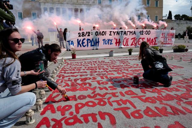 Students light flares as they hold a banner that reads “28/2 everyone to Syntagma, its ether their profits or our lives” while other students paint the names of the 57 killed in the train accident of Tempi outside the Greek parliament during a rally two days before the third anniversary of Tempi railway collision in February 2023, in Athens on February 26, 2026. Workers, school pupils, university students, and citizens across Greece are preparing to take to the streets on February 28 to mark three years since the Tempi rail disaster the country's worst rail tragedy, that left 57 dead. (Photo by Aggelos NAKKAS / AFP)