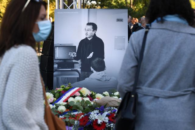 (FILES) Members of the public stand in front of a photograph depicting French history and geography teacher Samuel Paty as they attend a tribute ceremony in Eragny-sur-Oise, northwestern Paris, on October 16, 2021, held one year after Paty was beheaded by an extremist after showing his class cartoons of the Muslim Prophet Mohammed. On February 27, 2026, the prosecution requested that the sentences of the two initiators of a hate campaign against Samuel Paty be increased to 20 years' imprisonment on appeal. The campaign was launched before his beheading by a Chechen jihadist for showing caricatures of the Prophet during a lesson on freedom of expression. (Photo by Alain JOCARD / AFP)
