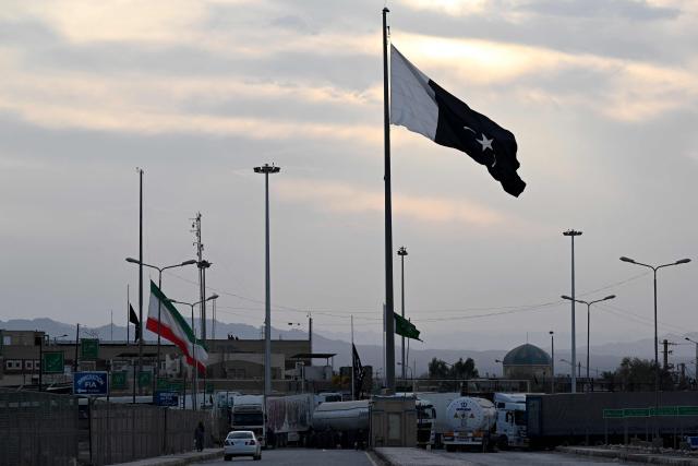 Cargo trucks cross into the Pakistan-Iran border as an Iranian national flag (L) flies at half-mast at Taftan, Balochistan province on March 1, 2026 after the death of Iran's supreme leader Ayatollah Ali Khamenei amid US-Israel strikes. (Photo by Banaras KHAN / AFP)