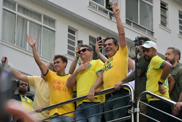 Brazilian Federal Deputy Nikolas Ferreira (L) and Senator and presidential candidate Flavio Bolsonaro (C) wave to supporters of Brazil's former president Jair Bolsonaro (2019-2022), sentenced to 23 years and 3 months for attempted coup, during a demonstration in Sao Paulo, Brazil, on March 1, 2026. (Photo by NELSON ALMEIDA / AFP)