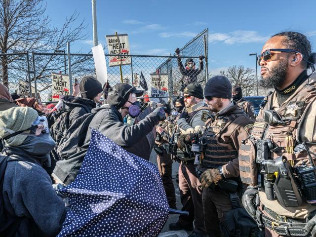 Law enforcement stands guard as protestors demonstate outside the Bishop Henry Whipple Federal Building during a protest opposing US Immigration and Customs Enforcement (ICE) operations, in Minneapolis, Minnesota, on March 1, 2026. The demonstration was organized in connection with a prayer camp established across the street at the sacred site of Mni Owe Sni (Coldwater Spring), where Indigenous organizers have been gathering in recent weeks to pray for families affected by immigration enforcement and to call for “ICE out” and “Land Back.” Indigenous leaders say immigration enforcement and detention echo historic patterns of displacement and incarceration experienced by Native communities. The camp at Mni Owe Sni — considered a sacred Dakota site — was established to center ceremony, prayer and solidarity with immigrant families. (Photo by Kerem YUCEL / AFP)