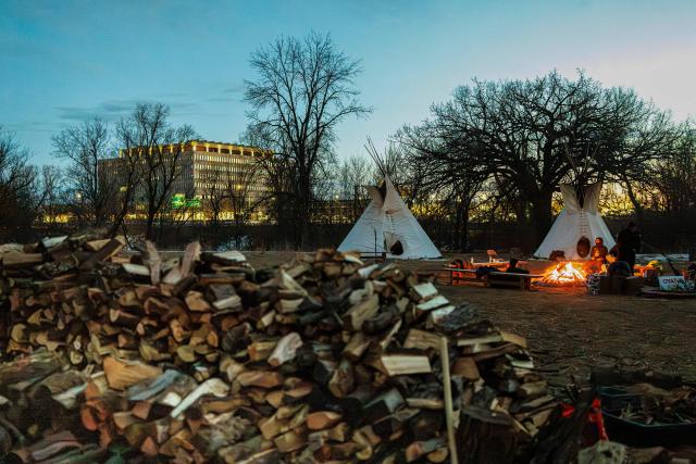 People gather around a bonfire at the Indigenous-led prayer camp across from the Bishop Henry Whipple Federal Building in Minneapolis, Minnesota, on March 1, 2026. The prayer camp was established at the sacred site of Mni Owe Sni (Coldwater Spring), where Indigenous organizers have been gathering in recent weeks to pray for families affected by immigration enforcement and to call for "ICE out" and "Land Back." Indigenous leaders say immigration enforcement and detention echo historic patterns of displacement and incarceration experienced by Native communities. (Photo by Kerem YUCEL / AFP)