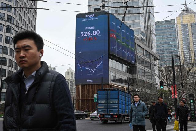 Pedestrians walk next to a screen showing the commodity futures for crude oil in the Jing'an district of Shanghai on March 2, 2026. Oil prices soared and stocks fell in Asia on March 2 after US-Israeli strikes on Iran sent investors fleeing the prospect of an extended conflict in the crude-rich Middle East. (Photo by Jade GAO / AFP)