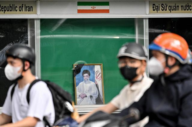 Motorcyclists ride past a portrait of Iran's late supreme leader Ayatollah Ali Khamenei at the Iranian Embassy in Hanoi on March 2, 2026, following his death amid US-Israel strikes. (Photo by Nhac NGUYEN / AFP)