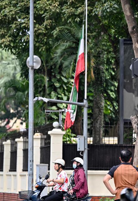 Women ride a motorbike past a flag of Iran at half-mast at the Iranian Embassy in Hanoi on March 2, 2026, following the death of Iran's supreme leader Ayatollah Ali Khamenei amid US-Israel strikes. (Photo by Nhac NGUYEN / AFP)