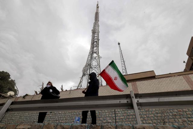 Iranians hold the national flag as they stand next to the destroyed state TV communications tower, next to Gandi Hospital, which was hit March 1, when a projectile struck during the ongoing joint US-Israeli military campaign on the capital Tehran on March 2, 2026. The United States and Israel launched strikes against Iran on February 28, killing Iran's supreme leader and top military leaders, prompting authorities to retaliate with strikes on Israel and across the Gulf. (Photo by ATTA KENARE / AFP)