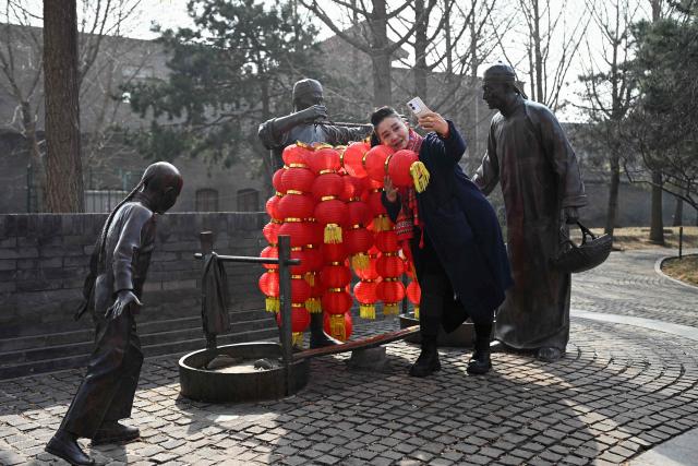 A woman takes a selfie with bronze statues decorated with lanterns at the entrance of an alley during the Lantern Festival in Beijing  on March 3, 2026. (Photo by WANG Zhao / AFP)