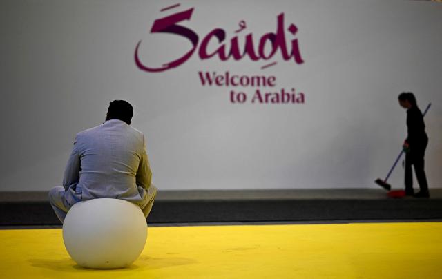A man sits at the booth of Saudi Arabia as a woman with a broom cleans the floor on the opening day of the ITB (Internationale Tourismus-Boerse) tourism fair in Berlin on March 3, 2026. Around 190 countries are represented at the platform for the global travel and tourism industry running from March 3 to 5, 2026. (Photo by Tobias SCHWARZ / AFP)