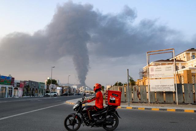 A delivery-person rides a motorcycle along a road as behind a tall smoke plume billows following an explosion in the Fujairah industrial zone on March 3, 2026. Iran's strikes on Gulf neighbours since February 28, following the US-Israeli attack, forced the UAE to shut its airspace, blindsiding travellers who thought they were headed to one of the region's safest holiday destinations. (Photo by Fadel SENNA / AFP)