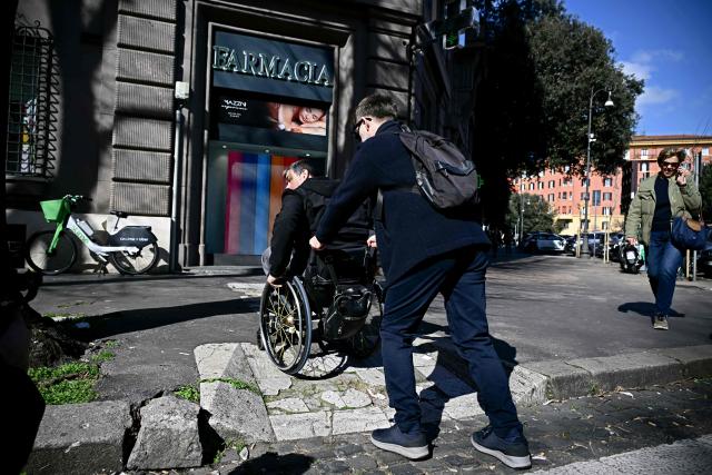 Italian lawyer Alessandro Bardini is assisted by a passerby while trying to use a damaged ramp to access the sidewalk after crossing a street in Rome on February 25, 2026. Paralympic organisers and Italy's government have invested tens of millions of euros in making the venues and areas around the Milan-Cortina Paralympic Games more accessible for people with disabilities. But the story is not the same across Italy, particularly in the capital Rome, known as much for its uneven roads and anarchic parking as for its ancient ruins. (Photo by Filippo MONTEFORTE / AFP)