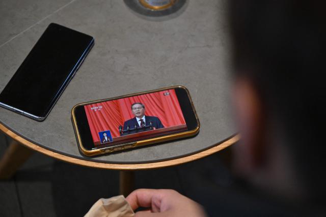 A man watches his phone showing a live broadcast of China's Premier Li Qiang delivering a speech during the opening session of the National People's Congress (NPC) at the Great Hall of the People, at a cafe in Beijing on March 5, 2026. (Photo by Pedro PARDO / AFP)