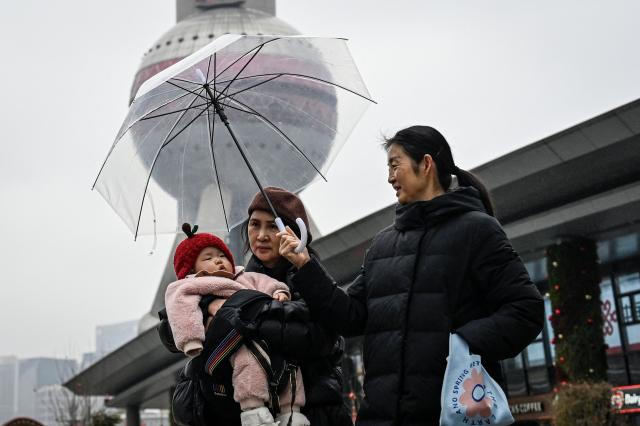 A woman holding a baby walks before the Oriental Pearl Tower as another woman holds an umbrella for them at the financial district of Lujiazui in Shanghai on March 5, 2026. (Photo by Jade GAO / AFP)