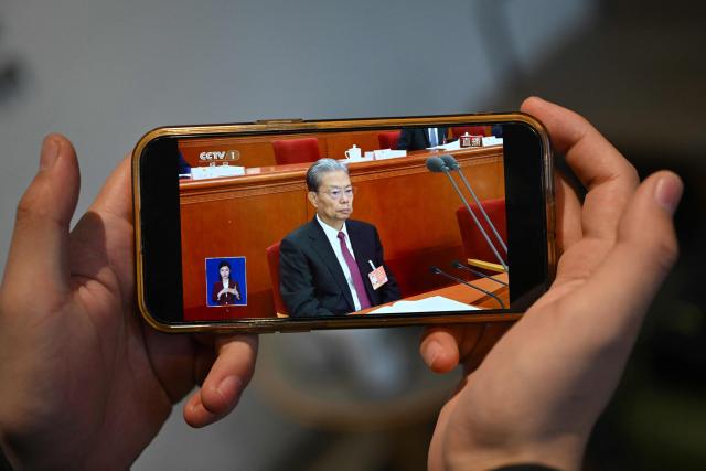 A man watches a live broadcast on his phone showing Zhao Leji, Chairman of the Standing Committee of China’s National People’s Congress (NPC), during the NPC's opening session at the Great Hall of the People, at a cafe in Beijing on March 5, 2026. (Photo by Pedro PARDO / AFP)