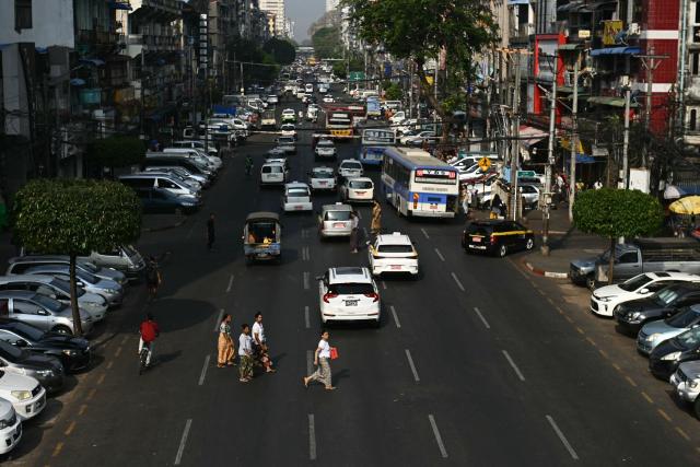 Pedestrians cross a road past vehicles on the first day of the implementation of an "odd-even" vehicle numbering system in Yangon on March 7, 2026. Myanmar's junta announced half of private vehicles will be ordered off the roads each day, based on licence plate numbers, in order to conserve fuel due to the war in the Middle East. Myanmar imports 90 percent of its fuel oil, according to 2024 figures, and has long suffered from a fragile energy supply chain owing to the civil war consuming the country since the military staged a coup five years ago. (Photo by Sai Aung MAIN / AFP)