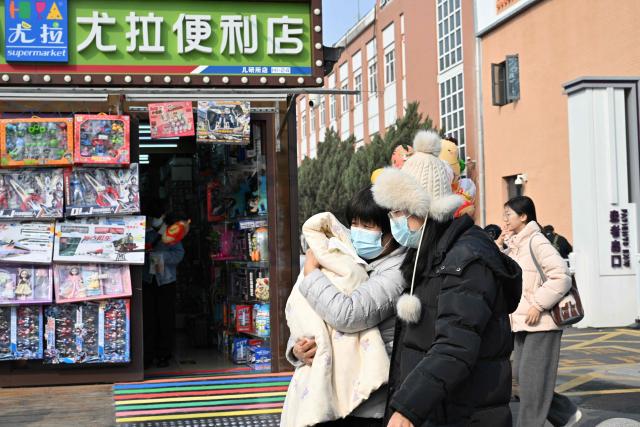People, including a woman holding a baby, walk past a shop selling toys beside a children's hospital in Beijing on March 7, 2026. (Photo by Adek BERRY / AFP)