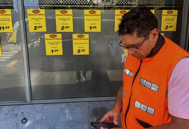 A man uses his mobile phone next to price signs on a supermarket window in Caracas on March 9, 2026. Venezuela closed 2025 with the highest inflation in the world: 475%, a brutal acceleration tied to the currency chaos triggered by the tightening of US sanctions against the ousted President Nicolas Maduro. (Photo by Juan BARRETO / AFP)