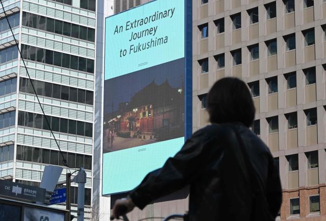 A woman rides a bicycle past a billboard showing a promotional video for tourism in Japan's Fukushima region and reading "An extraordinary journey to Fukushima", in downtown Seoul on March 10, 2026. A tourism promotion video for Japan's tsunami-hit Fukushima region is being shown on billboards across the South Korean capital Seoul for the first time since the 2011 nuclear accident, a Japanese official said on March 10. (Photo by Jung Yeon-je / AFP)