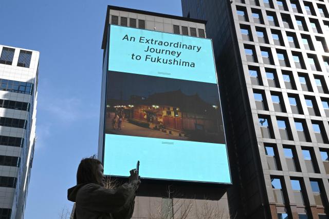 A woman uses her smartphone in front of a billboard showing a promotional video for tourism in Japan's Fukushima region and reading "An extraordinary journey to Fukushima", in downtown Seoul on March 10, 2026. A tourism promotion video for Japan's tsunami-hit Fukushima region is being shown on billboards across the South Korean capital Seoul for the first time since the 2011 nuclear accident, a Japanese official said on March 10. (Photo by Jung Yeon-je / AFP)