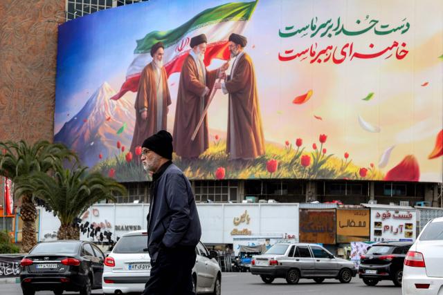 TOPSHOT - A man walks near a banner displayed at Valiasr Square in central Tehran on March 10, 2026, depicting Iran's late supreme leader Ayatollah Ruhollah Khomeini (L) watching as his successor the late Ayatollah Ali Khamenei (C) hands over a national flag to his son and new supreme leader Mojtaba Khamenei (R). Iran marked the appointment of Ayatollah Mojtaba Khamenei to replace his father as its supreme leader with a new barrage of missiles against Israel and the Gulf states on March 9, as the Middle East war sent oil prices soaring. (Photo by AFP) / Attention editors: AFP covers the war in the Middle East through its extensive regional network, including bureaus in Tehran, Jerusalem, and several neighboring countries. Since the start of the conflict, journalists have been working under increasingly restrictive conditions. Authorities in several countries have limited reporters' movements, photo and live video coverage from sensitive locations. Some governments and armed groups have banned images of missile or drone strikes and other security-related sites. / 
