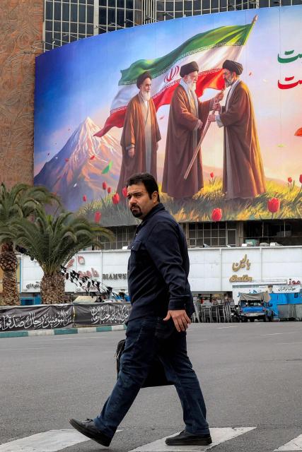 A man walks near a banner displayed at Valiasr Square in central Tehran on March 10, 2026, depicting Iran's late supreme leader Ayatollah Ruhollah Khomeini (L) watching as his successor the late Ayatollah Ali Khamenei (C) hands over a national flag to his son and new supreme leader Mojtaba Khamenei. Iran marked the appointment of Ayatollah Mojtaba Khamenei to replace his father as its supreme leader with a new barrage of missiles against Israel and the Gulf states on March 9, as the Middle East war sent oil prices soaring. (Photo by AFP) / Attention editors: AFP covers the war in the Middle East through its extensive regional network, including bureaus in Tehran, Jerusalem, and several neighboring countries. Since the start of the conflict, journalists have been working under increasingly restrictive conditions. Authorities in several countries have limited reporters' movements, photo and live video coverage from sensitive locations. Some governments and armed groups have banned images of missile or drone strikes and other security-related sites. / 