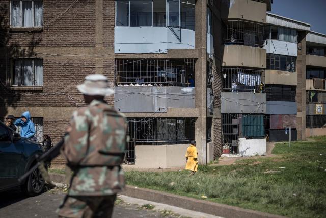 A resident (R) strolls near an apartment block as unseen South Africa Police Service (SAPS) officers and South African National Defence Force (SANDF) soldiers conduct a house search in Westbury, Johannesburg, on March 11, 2026. Soldiers moved into gang-violence hotspots in Johannesburg on March 11, 2026 to support police efforts to tackle rampant crime, described by President Cyril Ramaphosa as one of the biggest threats facing South Africa. (Photo by MARCO LONGARI / AFP)