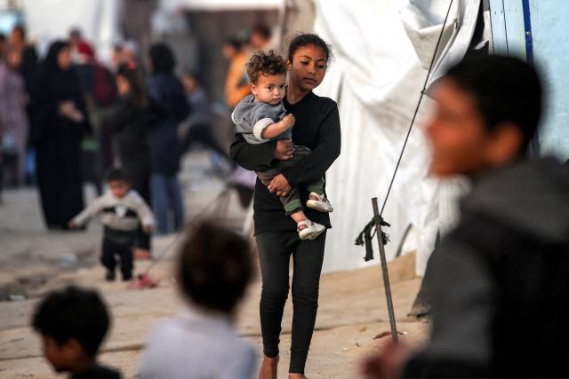 TOPSHOT - A girl walks carrying a child past tents sheltering Palestinians displaced by conflict in Nuseirat in the central Gaza Strip on March 11, 2026. (Photo by Eyad Baba / AFP)