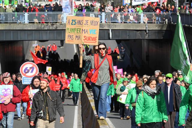 A protester holds a sign reading "At 25 years, I want to plan my life, not my survival" during a demonstration during a national day of action against the austerity plan of the federal Arizona government, in Brussels on March 12, 2026. Belgium will be running at a slower pace on March 12, 2026 due to a call for a strike by the country’s three main trade unions, resulting in the suspension of all traffic at Charleroi Airport and the cancellation of departing flights from Brussels-Zaventem. (Photo by Nicolas TUCAT / AFP)