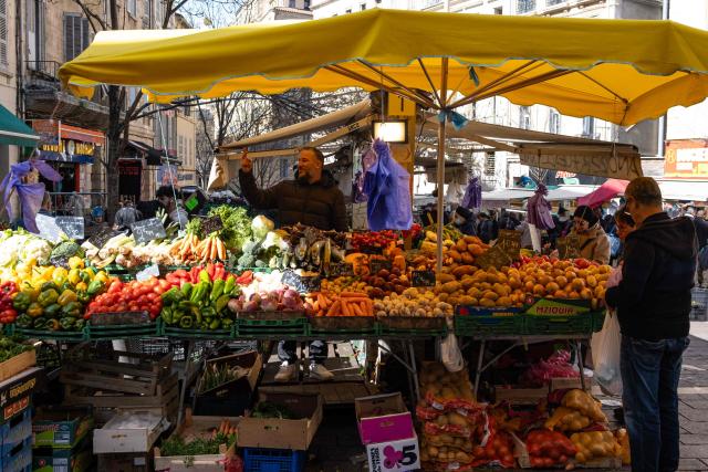 A stall holder gestures as he stands behind his vegetable stall at a market in Marseille, southern France, on March 12, 2026. (Photo by Elodie CLEMENT / AFP)