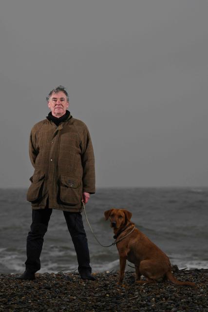 This photo taken on February 3, 2026 shows Thorpeness resident Nicholas Millor with his dog Demelza posing for a photograph in Thorpeness, Suffolk. In an English seaside village, researchers discuss options for relocating a graveyard threatened with slipping into the sea, or moving back a car park perilously close to a cliff edge. The team from the Coastwise project have been granted over 15 million pounds (20 million USD) in government funding to adapt the coastline in North Norfolk, eastern England, to accelerating erosion worsened by climate change. (Photo by Ben STANSALL / AFP) / TO GO WITH Britain-climate-environment,FOCUS by Akshata KAPOOR