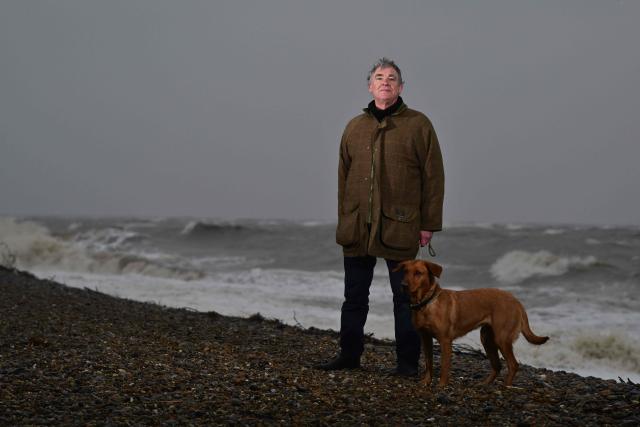 This photo taken on February 3, 2026 shows Thorpeness resident Nicholas Millor with his dog Demelza posing for a photograph in Thorpeness, Suffolk. In an English seaside village, researchers discuss options for relocating a graveyard threatened with slipping into the sea, or moving back a car park perilously close to a cliff edge. The team from the Coastwise project have been granted over 15 million pounds (20 million USD) in government funding to adapt the coastline in North Norfolk, eastern England, to accelerating erosion worsened by climate change. (Photo by Ben STANSALL / AFP) / TO GO WITH Britain-climate-environment,FOCUS by Akshata KAPOOR