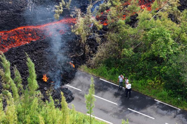 This aerial photograph shows a police officer and two pedestrians standing next to a lava flow making its way through a forested area crossing the RN2 road near Sainte-Rose close to the Piton de la Fournaise volcano in the southeast of the French overseas island of Reunion on March 13, 2026. Two lava flows spewed out by Piton de la Fournaise, a volcano that has been erupting for a month on Réunion Island, have cut the national road linking the south to the east of the islandsomething that has not happened in nearly 20 years, the prefecture said on Friday. (Photo by Richard BOUHET / AFP)