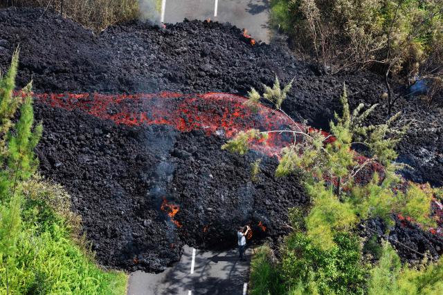 This aerial photograph shows a person standing next to a lava flow making its way through a forested area crossing the RN2 road near Sainte-Rose close to the Piton de la Fournaise volcano in the southeast of the French overseas island of Reunion on March 13, 2026. Two lava flows spewed out by Piton de la Fournaise, a volcano that has been erupting for a month on Rйunion Island, have cut the national road linking the south to the east of the island—something that has not happened in nearly 20 years, the prefecture said on Friday. (Photo by Richard BOUHET / AFP)