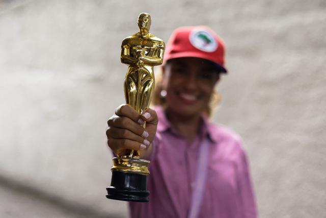 A tourist poses for a photo holding a replica of the Oscar statue during a guided tour through the scenarios of the film "O Agente Secreto" (The Secret Agent) at the city center of Recife, Pernambuco state, Brazil, on February 13, 2026. The northeastern Brazilian city of Recife, where the Oscar-nominated movie "The Secret Agent" is set, is revelling in its moment in the limelight as tourists flock to film locations and a classic local shirt flies off the shelves. (Photo by BRENDA ALCANTARA / AFP)