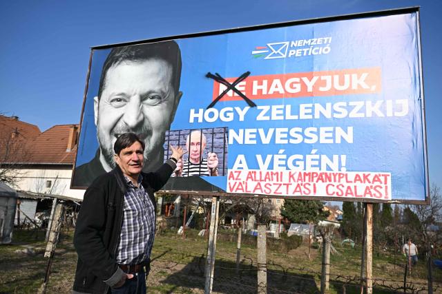 Human rights activist Peter Heindl stands next to a “corrected” government billboard showing Ukrainian President Volodymyr Zelensky and the slogan “Let’s not let Zelensky have the last laugh”, in Pecs, Hungary on March 13, 2026. The word “not” was crossed out, and a photo of Vladimir Putin with a sign saying “the state campaign billboard is election fraud” was added. Heindl has been modifying billboards for years, arguing that the Orban government uses state resources to skew elections. (Photo by Attila KISBENEDEK / AFP)
