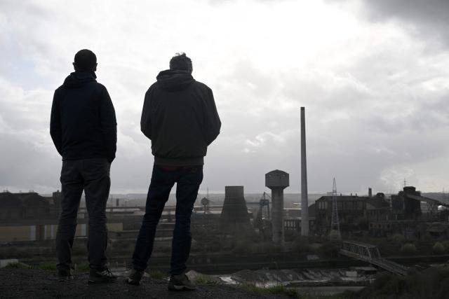 Tourists stand as they look at abandoned steel plants in Charleroi, southern Belgium, on March 11, 2026. Once a major industrial city of the coal industry, Charleroi still bears the marks of a working-class past now lying fallow. As they venture through its abandoned factories, urban explorers (urbex) end up telling the story of a system as much as of a pastime: that of a capitalism which pours investment into a place, exploits it, then withdraws, leaving behind nothing but ruins to explore. (Photo by Nicolas TUCAT / AFP)