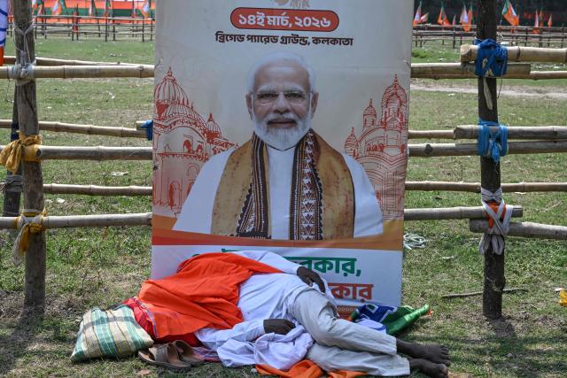 A Bharatiya Janata Party (BJP) supporter takes a nap near the poster of India's Prime Minister Narendra Modi during a mass rally in Kolkata on March 14, 2026. (Photo by Dibyangshu SARKAR / AFP)