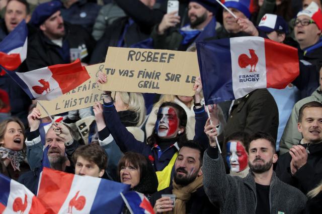 A Frrnch supporter holds a placard that reads "Rosbeef You're going to end up on the grill" as they cheer their team on from the stands ahead of the Six Nations international rugby union match between France and England at the Stade de France, in Saint-Denis, north of Paris, on March 14, 2026. (Photo by Thomas SAMSON / AFP)