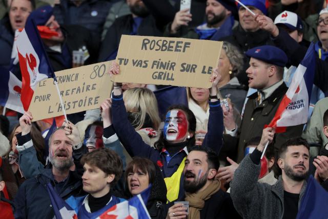 A Frrnch supporter holds a placard that reads "Rosbeef You're going to end up on the grill" as they cheer their team on from the stands ahead of the Six Nations international rugby union match between France and England at the Stade de France, in Saint-Denis, north of Paris, on March 14, 2026. (Photo by Thomas SAMSON / AFP)