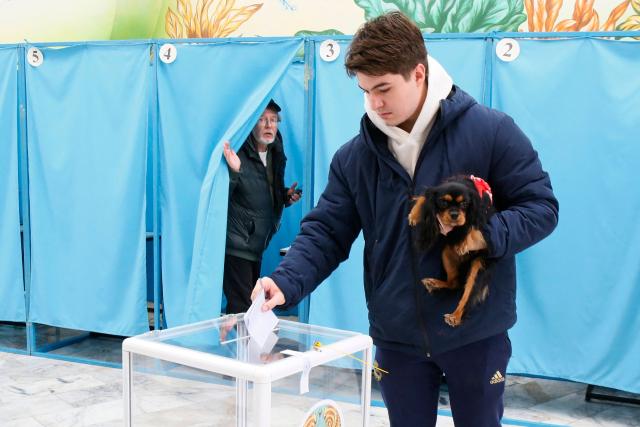 A man holding a dog casts his ballot as another voter leaves a voting booth at a polling station during the constitutional referendum in Almaty on March 15, 2026. (Photo by Ruslan PRYANIKOV / AFP)