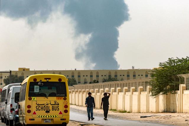 Two men walk past parked vehicles along a road as a smoke plume rises from an ongoing fire at Dubai International Airport in Dubai on March 16, 2026. Flights were gradually resuming at Dubai airport on March 16, previously the world's busiest for international flights, the airport operator said, after a "drone-related incident" sparked a fuel tank fire nearby, as Iran kept up its Gulf attacks. (Photo by AFP) / 