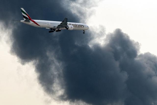 TOPSHOT - An Emirates Airbus A350 aircraft prepares for landing as a smoke plume rises from an ongoing fire at Dubai International Airport in Dubai on March 16, 2026. Flights were gradually resuming at Dubai airport on March 16, previously the world's busiest for international flights, the airport operator said, after a "drone-related incident" sparked a fuel tank fire nearby, as Iran kept up its Gulf attacks. (Photo by AFP) / 