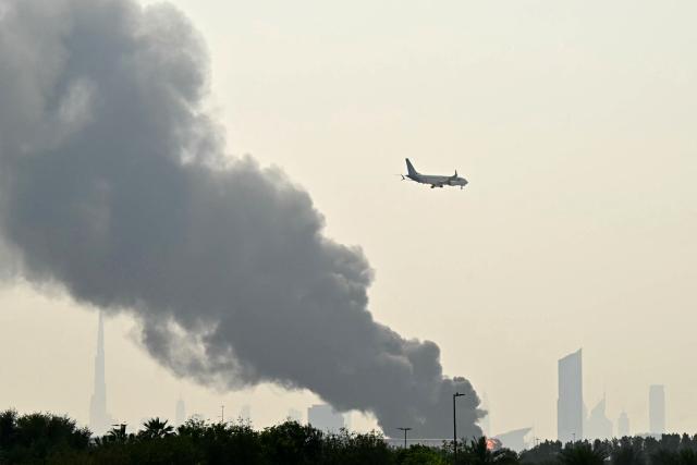 An Emirates aircraft flies past plumes of smoke from an ongoing fire near Dubai International Airport in Dubai on March 16, 2026. Missiles and drone attacks hit across the UAE, with a drone-related incident sparking a fuel tank fire near Dubai airport that disrupted travel, while a missile killed a civilian in Abu Dhabi. (Photo by AFP) / 