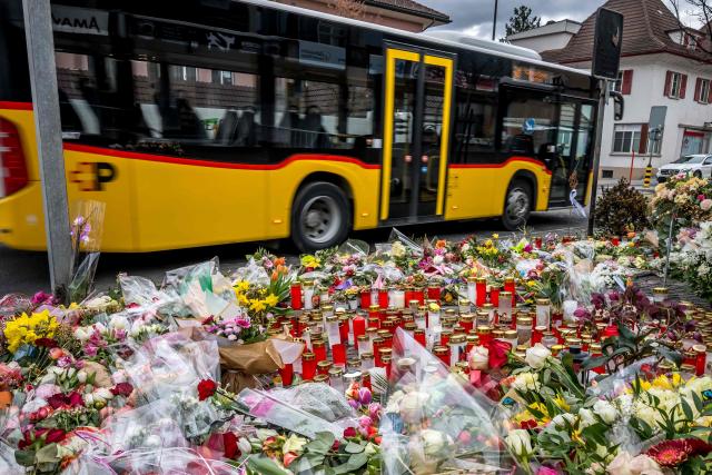 A PostBus passes flowers and candles at the site where a bus from the same public transport company caught fire after a man described as "disturbed" set himself alight, killing at least six people and injuring five others, in Kerzers, western Switzerland, on March 16, 2026. (Photo by Fabrice COFFRINI / AFP)