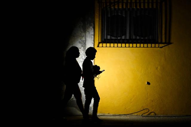 A woman holds a flashlight while walking with a man on a street during a blackout in Havana on March 16, 2026. Cuba suffered a widespread power cut on March 16, 2026, according to the national electricity company, against the backdrop of a severe crisis on the island caused by the US energy blockade. (Photo by Yamil LAGE / AFP)